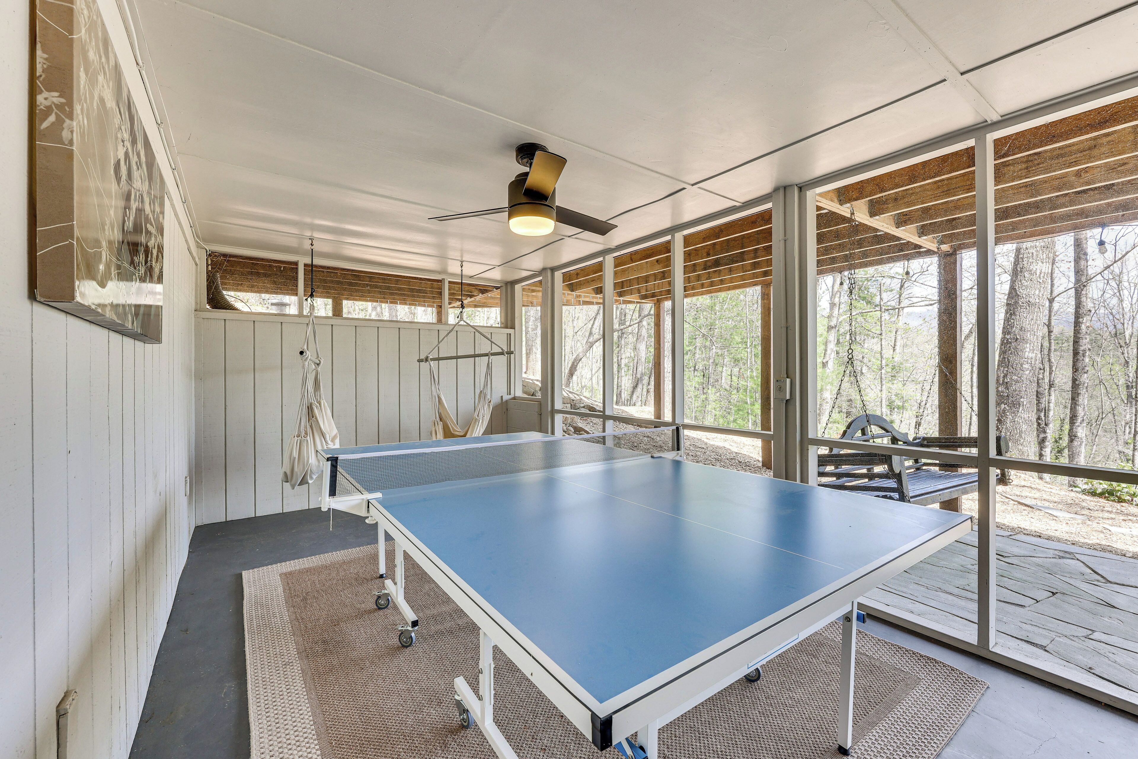 Une table de ping-pong installée dans un espace abrité, doté de grandes fenêtres, d’un ventilateur de plafond et offrant une vue sur les arbres.