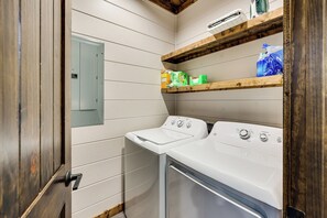 A laundry room with two white washers and dryers, wooden shelves above, and a door with a handle. - Two King Suites, Private Hot Tub & Treetop Deck (Broken Bow)