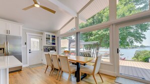 A modern kitchen with white cabinets, a stainless steel refrigerator, and a dining area with a wooden table and chairs. Large windows offer a view of a deck and a body of water. - 'commodore Bay Waterfront Home on Lake Norman! (Mooresville)