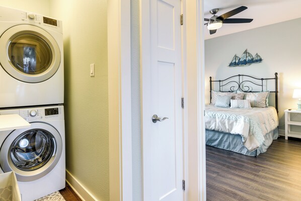 Un cuarto de lavado con lavadora y secadora frontales, una puerta que da a un dormitorio con cama, mesita de noche y ventilador de techo.