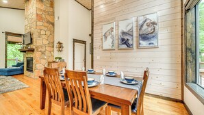 A dining area with a stone fireplace, wooden dining table, and chairs. There are abstract paintings on the wall and a large window providing a view of greenery.