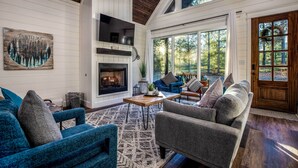 A spacious living room with a fireplace, blue and gray furniture, and large windows offering a view of the outdoors. - 'painted Sky' Modern Broken Bow Cabin w/ Hot Tub! (Broken Bow)