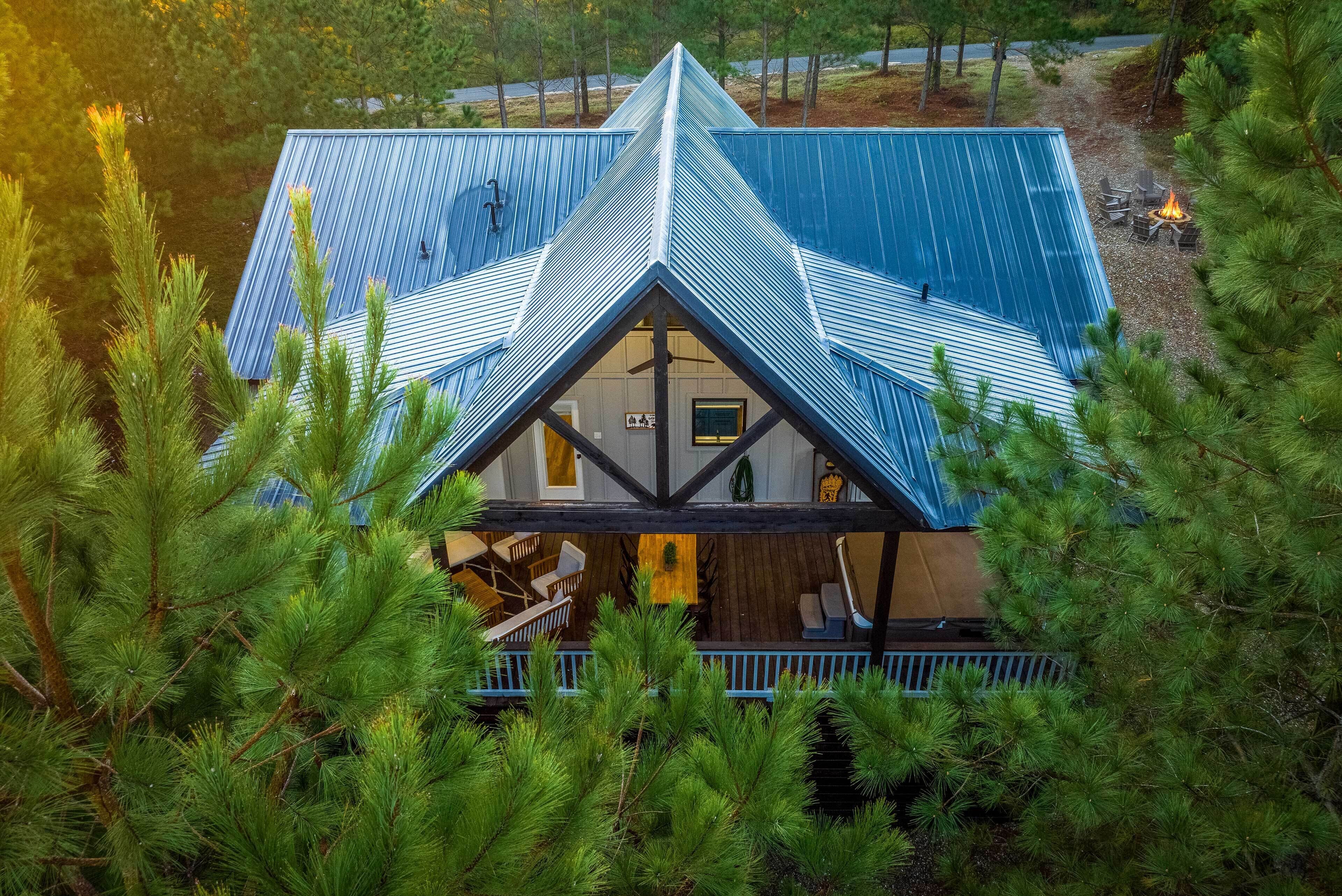 A cabin with a triangular roof, surrounded by pine trees, featuring a deck with outdoor seating and a fire pit.