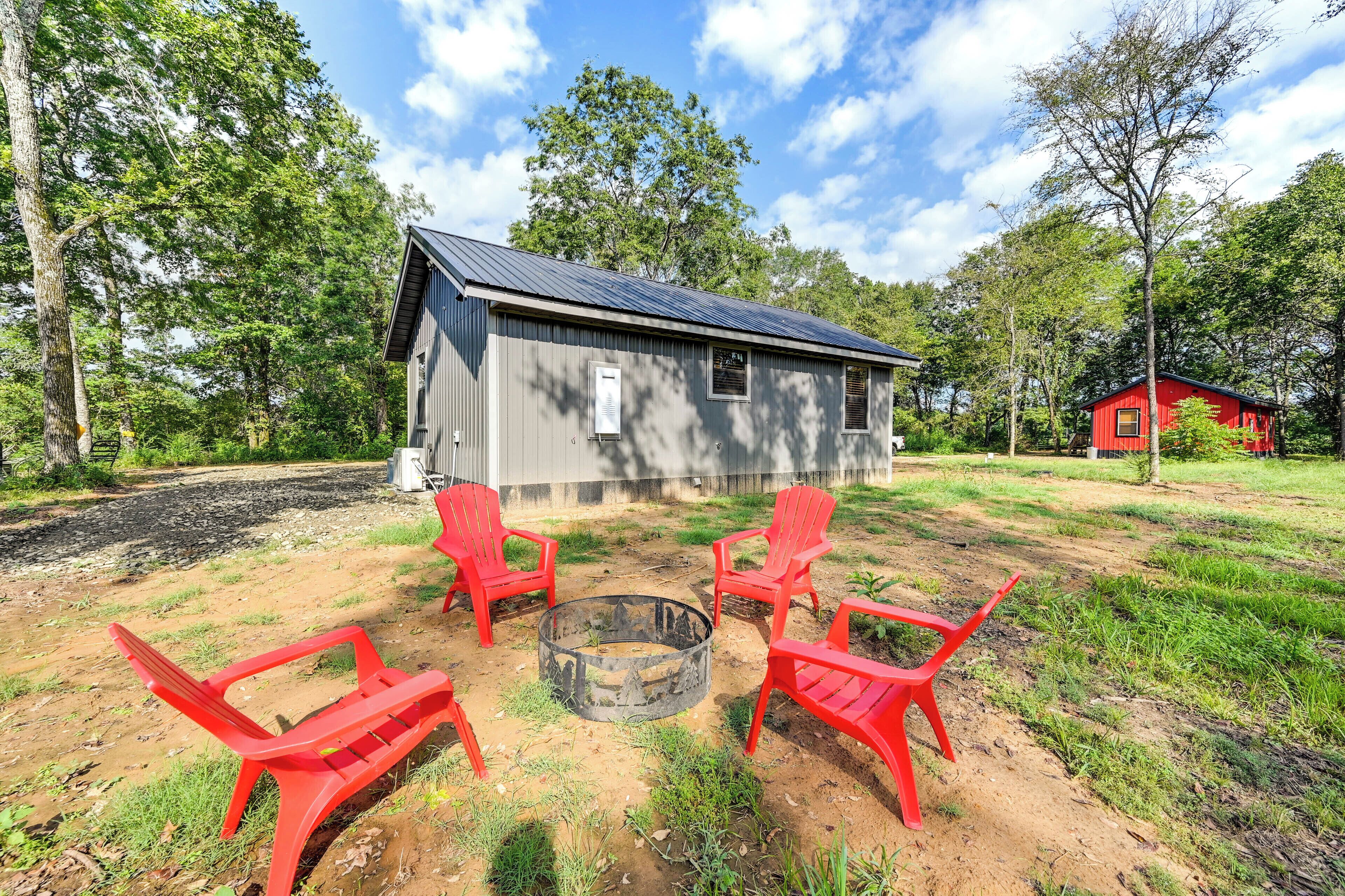 A rustic cabin with a metal roof, surrounded by a grassy area with red plastic chairs and a fire pit.