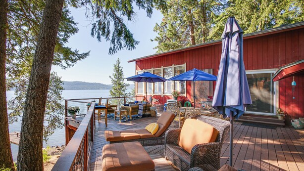 A deck with wicker furniture, blue umbrellas, and a red building with a glass door. - Lakefront Bliss: Coeur D'alene Cabin w/ Dock! (Coeur d'Alene)