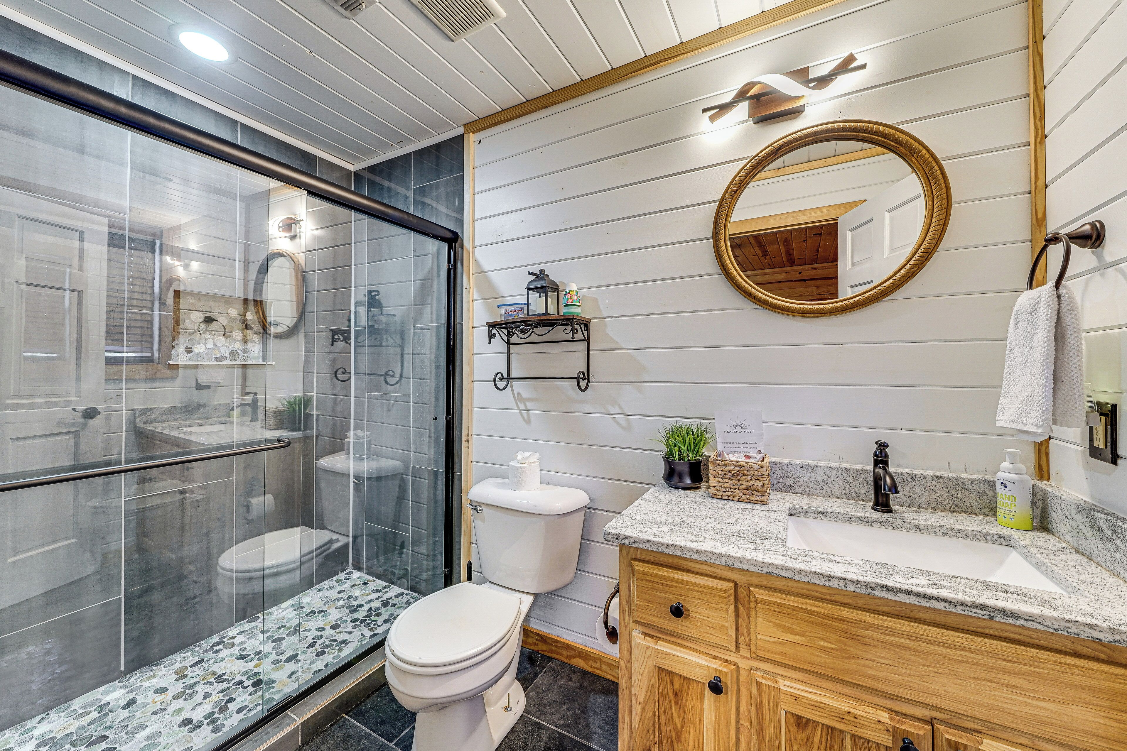 A modern bathroom with a glass shower enclosure, a white toilet, a granite countertop sink, wooden cabinetry, and a round mirror.