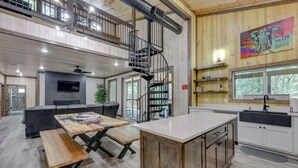 A spacious interior with a wooden floor, a kitchen with white cabinets, a dining area with a wooden table and benches, a black staircase, and a large window with a view of trees. - Family-friendly Broken Bow Home w/ Deck & Grill! (Broken Bow)