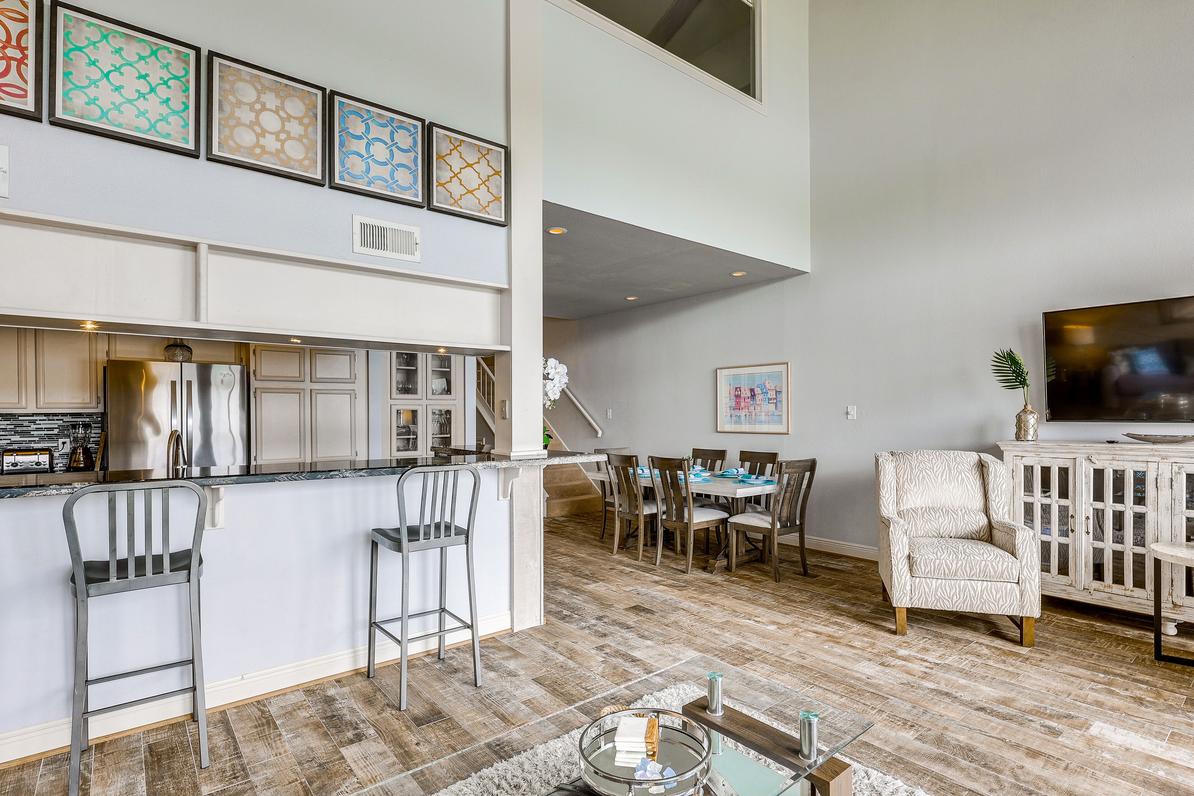 A modern kitchen with bar stools, a dining area with a table and chairs, and a living room with a TV and patterned armchair.