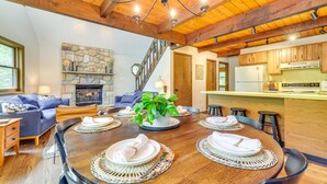 A cozy living room with a stone fireplace, a dining table set for four, and a kitchen area with wooden cabinets and a countertop.