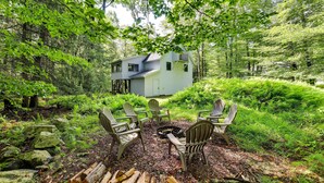 A cabin surrounded by dense forest, with a fire pit and Adirondack chairs in the foreground.