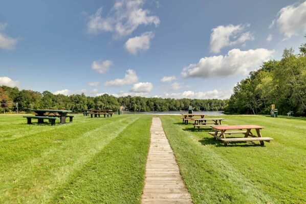 A picnic area with a wooden path leading to a lake, surrounded by green grass and trees.