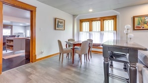A dining area with a wooden table and chairs, a kitchen in the background, and a window with curtains. - Stillwater Home - Walk to Boone Pickens Stadium! (Stillwater)