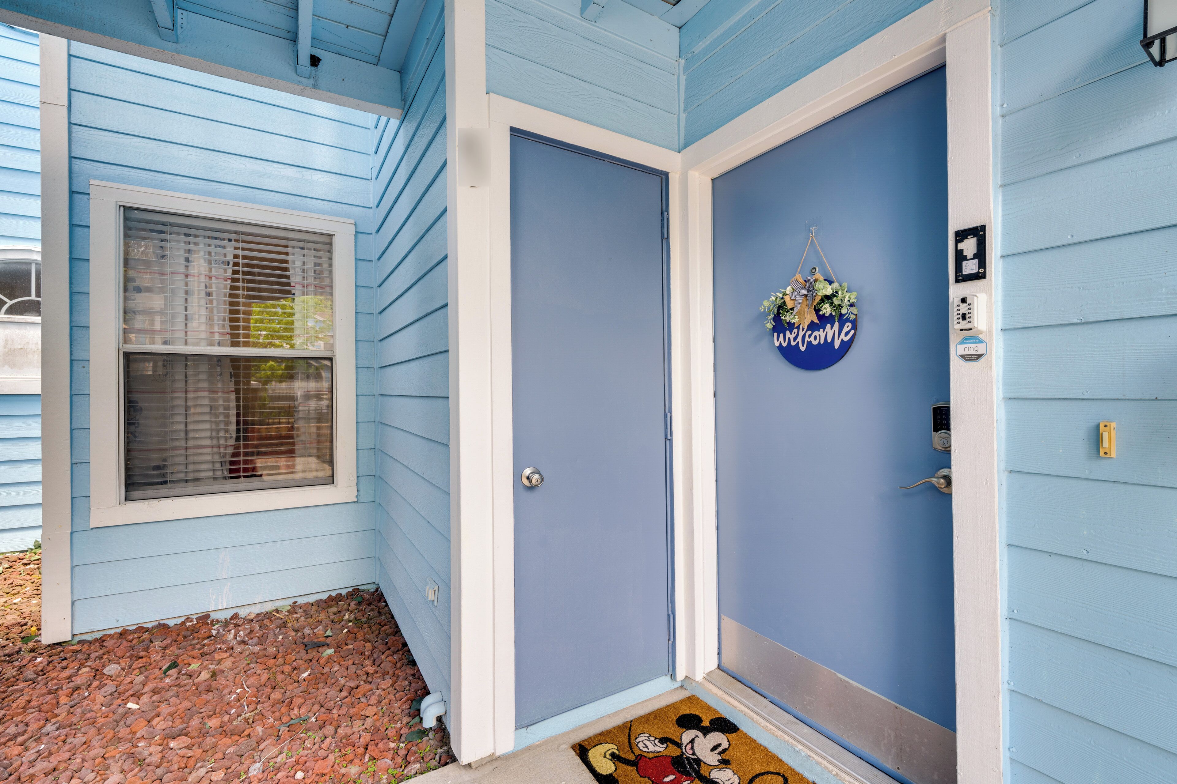 A house with a blue exterior, a front door with a 'welcome' sign, a window with blinds, and a doormat with Mickey Mouse.