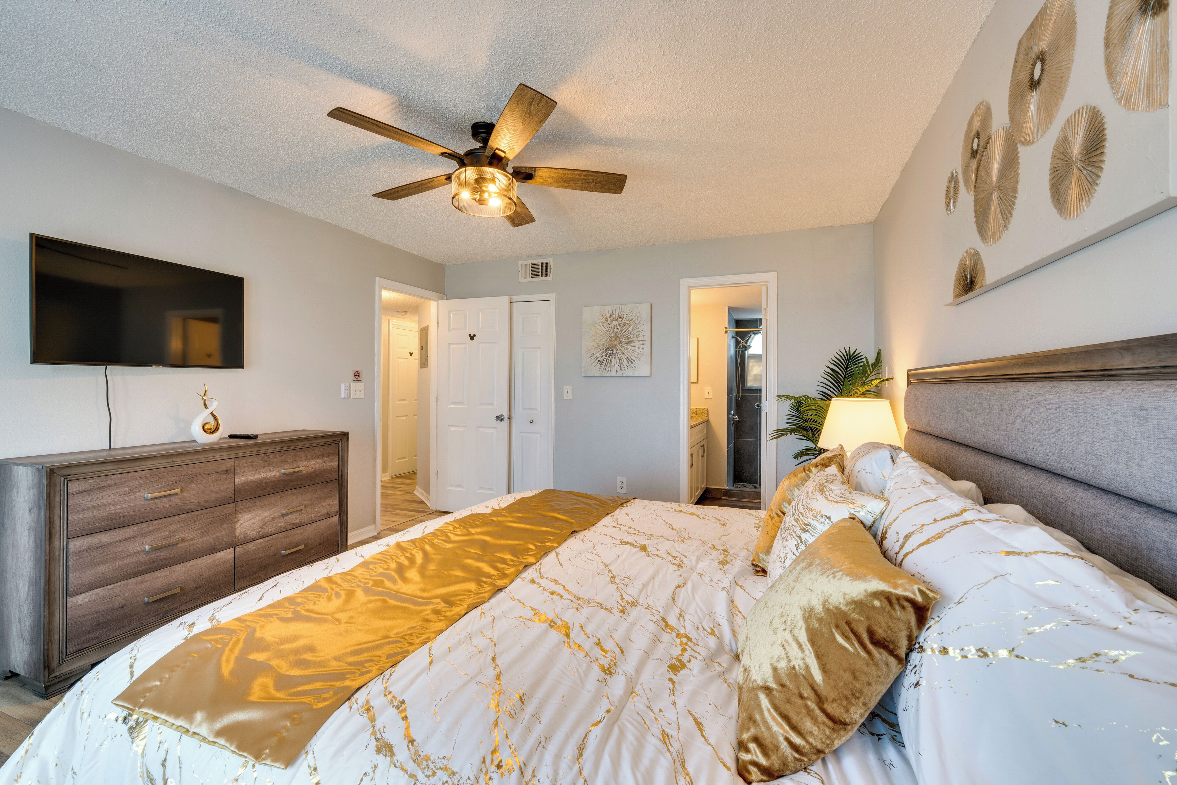 A bedroom with a bed, a ceiling fan, a TV, a dresser, and a door leading to another room.