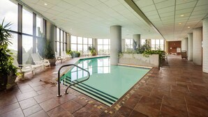 Une piscine intérieure aux parois en verre transparent, entourée de chaises longues blanches et de plantes en pot.