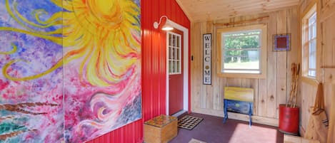 Interior d'una cabana de fusta amb un quadre gran i colorit, un cartell de benvinguda i una banca de fusta rústica.