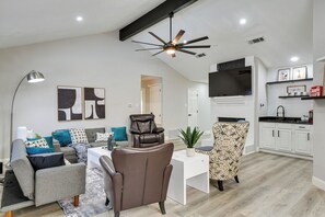 A modern living room with a sofa, armchair, and coffee table, a dining area with a white table, and a kitchen with white cabinets and a black countertop.