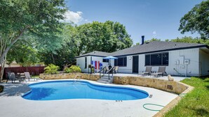 Ein Schwimmbecken mit Steinbord, eine überdachte Terrasse mit Stühlen und ein Haus im Hintergrund.