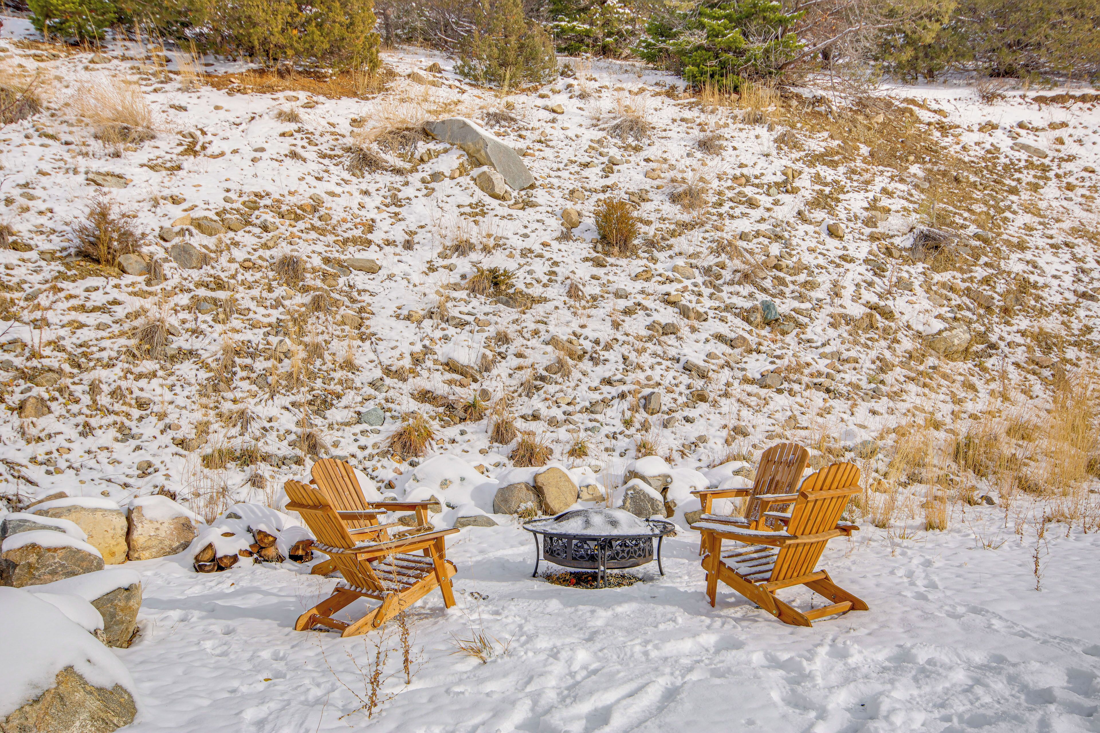 Twee houten Adirondack stoelen gericht op een metalen vuurkuil in een besneeuwde omgeving met rotsen en schaarse begroeiing.