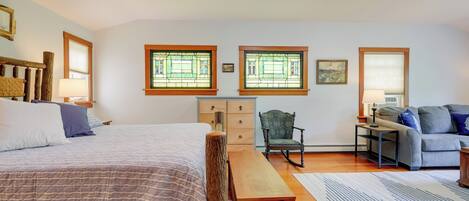 Une chambre à coucher avec un grand lit, un banc en bois, un fauteuil à bascule, un canapé et deux fenêtres ornées de vitraux.