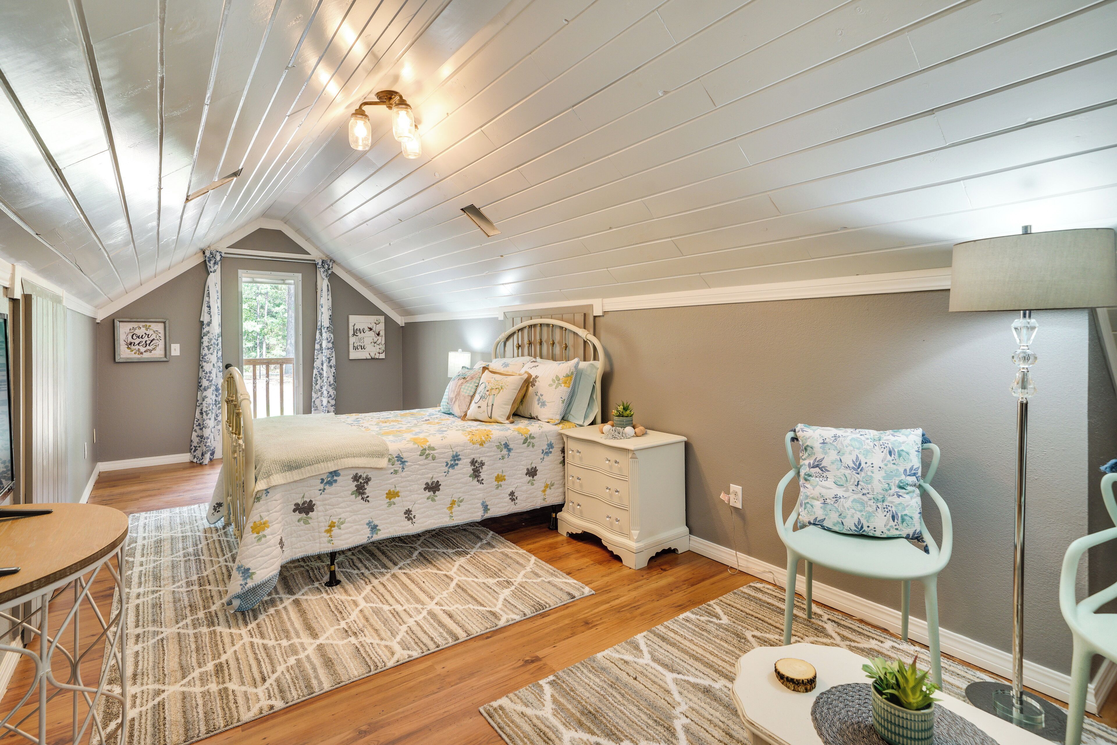 A bedroom with a sloped ceiling, a bed with floral bedding, a dresser, a chair, a lamp, and a small plant.