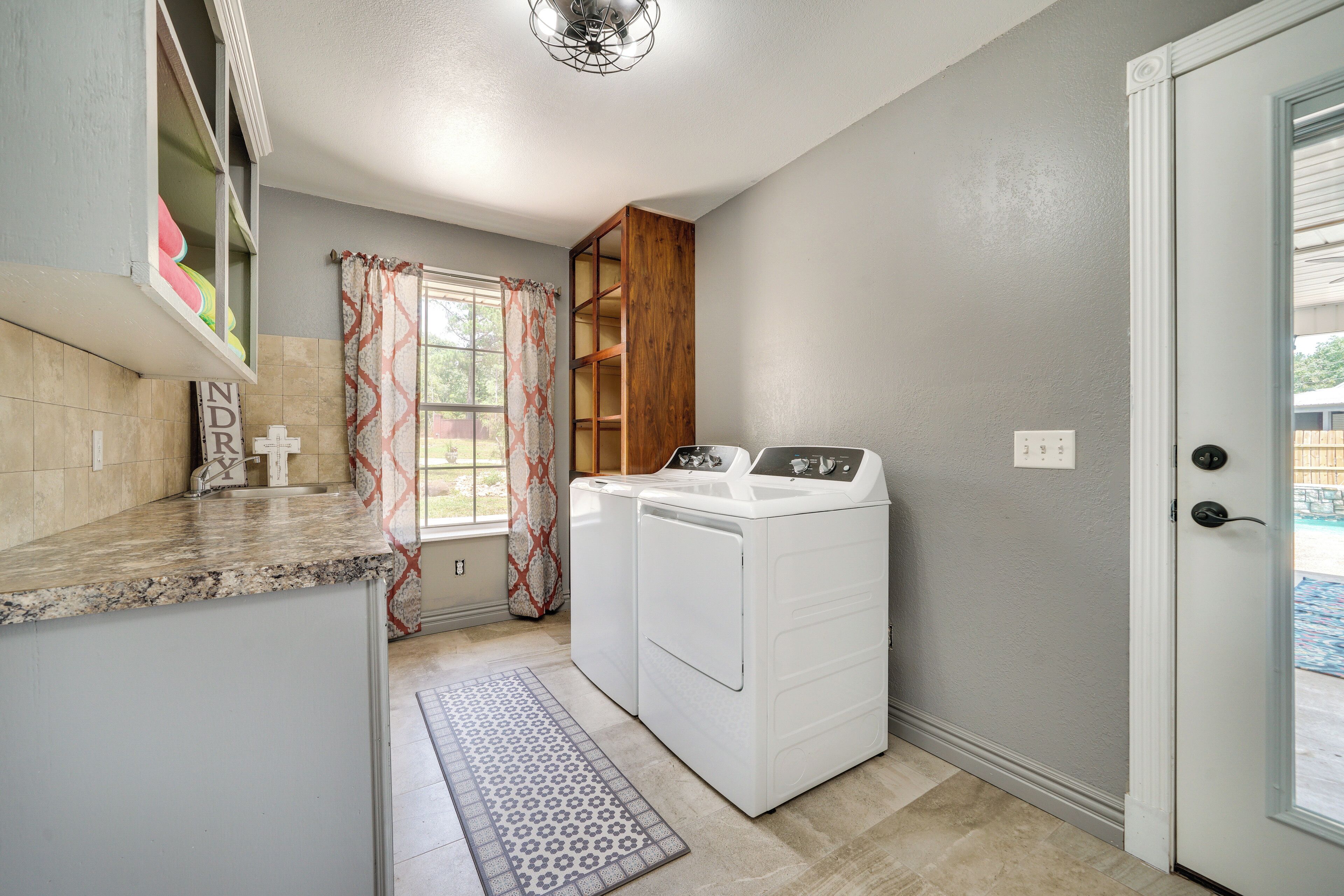 A laundry room with a washing machine, a drying rack, and a window with curtains.