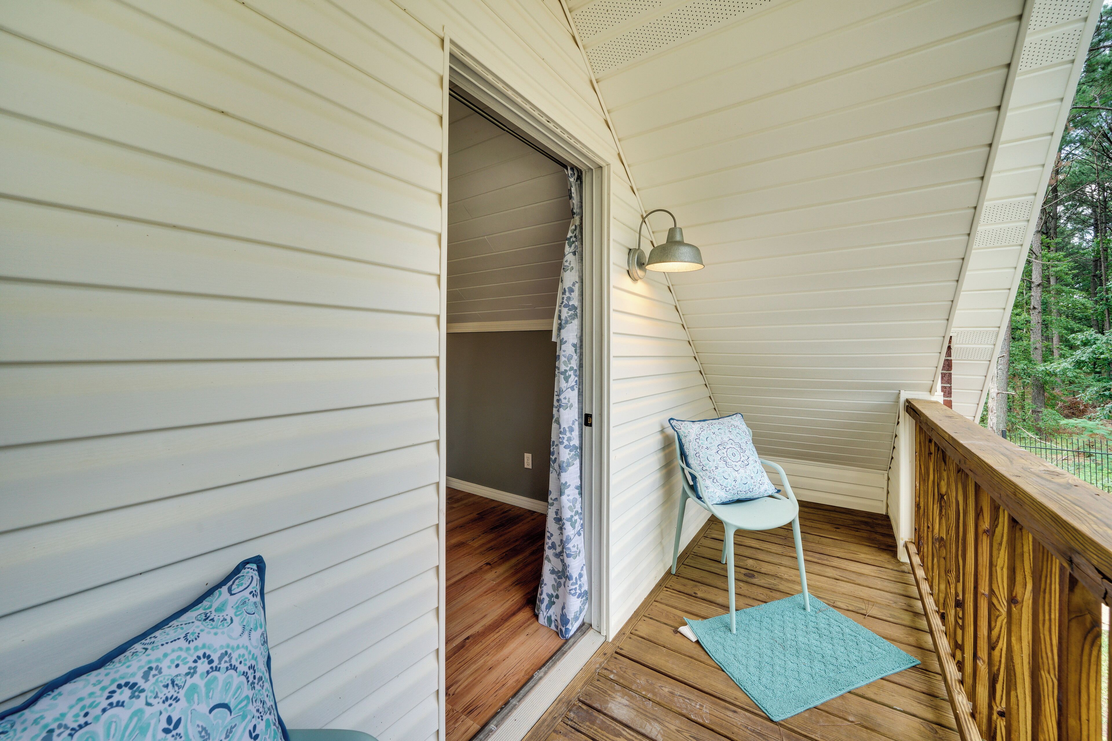 Une véranda couverte avec un plancher en bois, une chaise blanche avec un coussin bleu, une petite table et une lampe fixée au mur.
