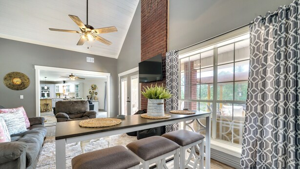 A spacious living area with a high ceiling fan, a dining table with stools, a sofa, and a large window with patterned curtains. - Broken Bow Vacation Rental w/ Pool & Hot Tub! (Broken Bow)