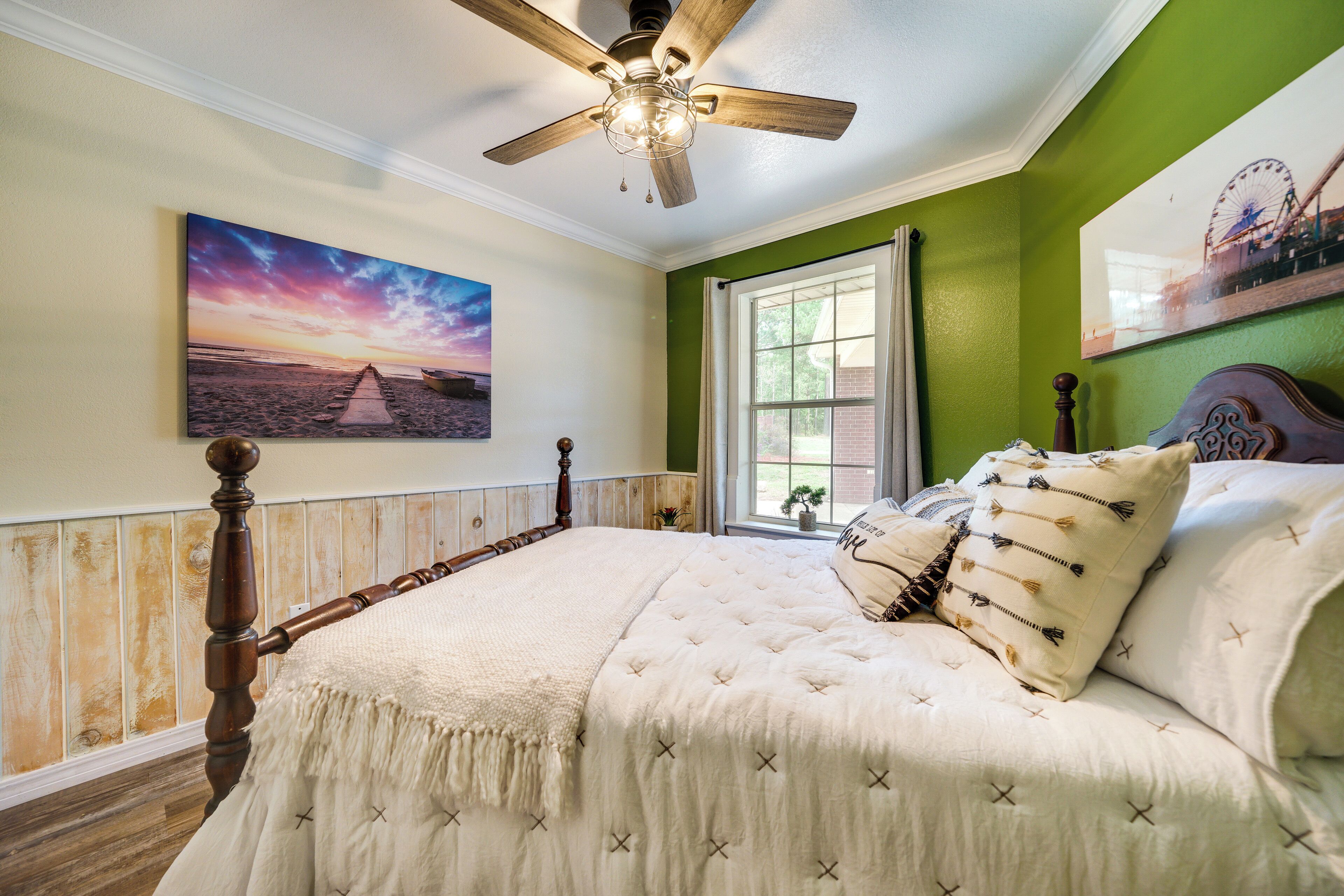 Une chambre avec un lit en bois, un ventilateur de plafond, une grande fenêtre donnant sur un beau paysage et un tableau accroché au mur.
