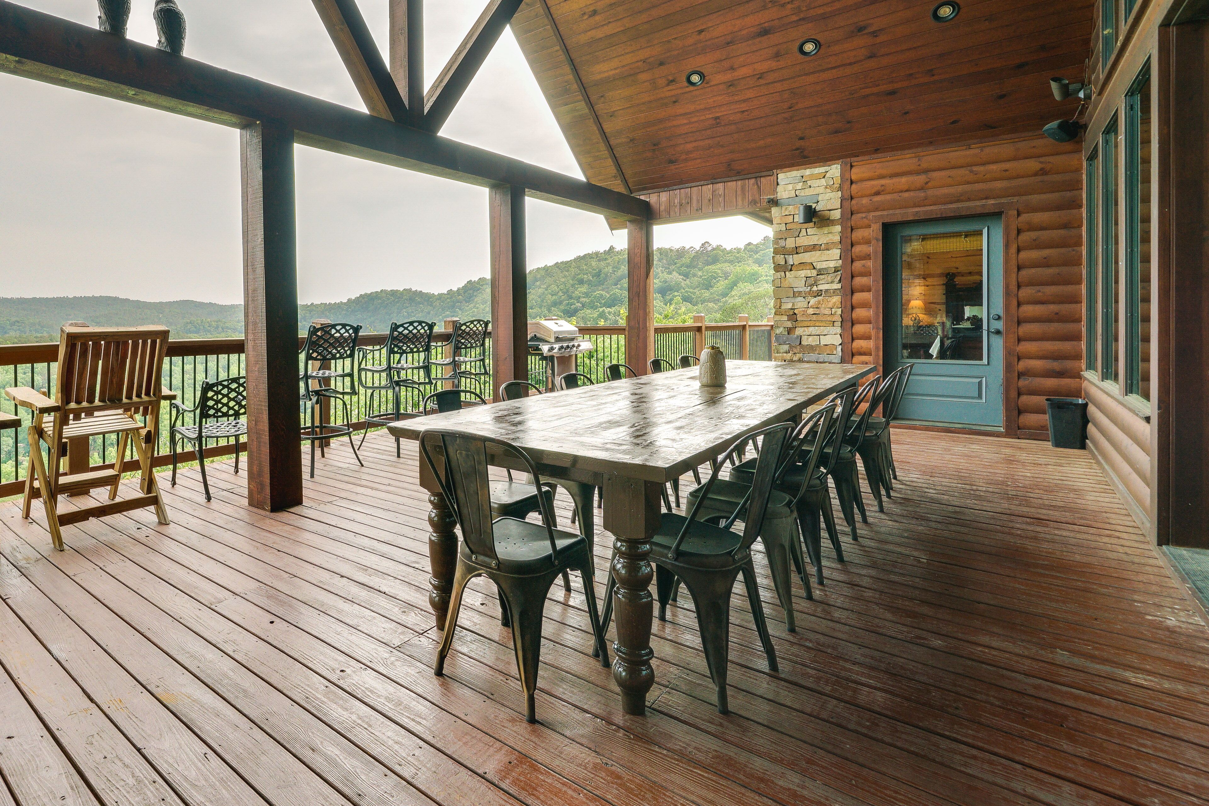 A covered outdoor deck with a wooden table and chairs, a stone wall, and a view of a forested hill.