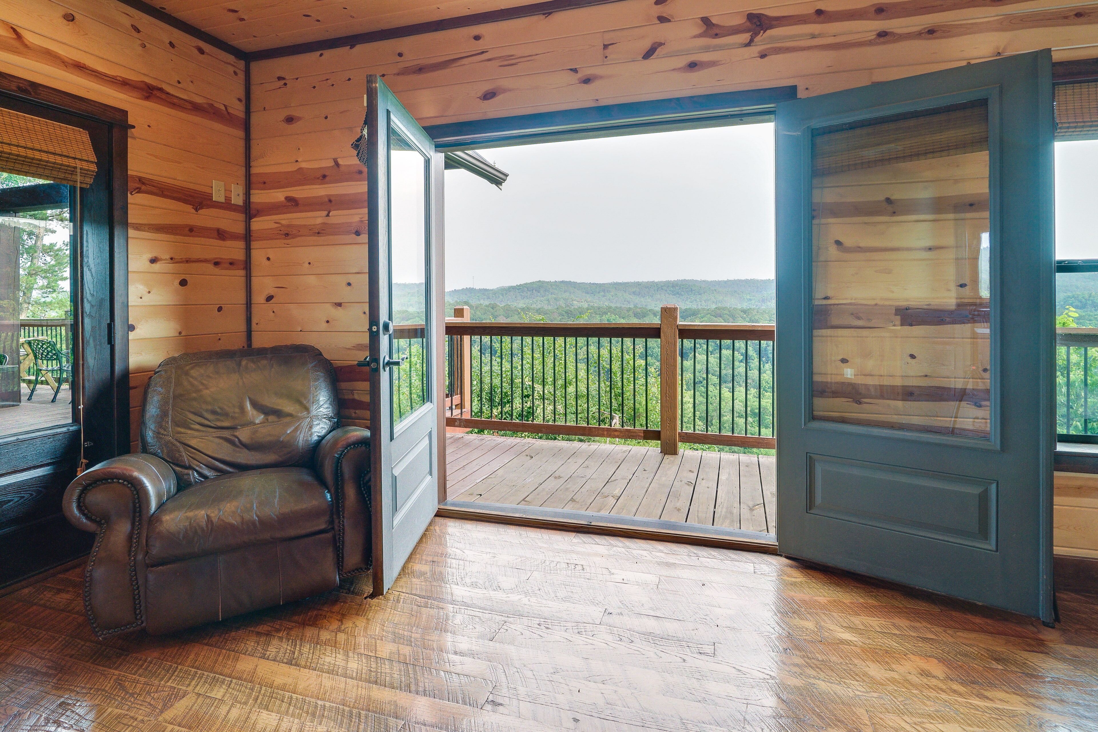 A wooden interior with a leather chair, a balcony with a wooden deck, and a view of green hills.