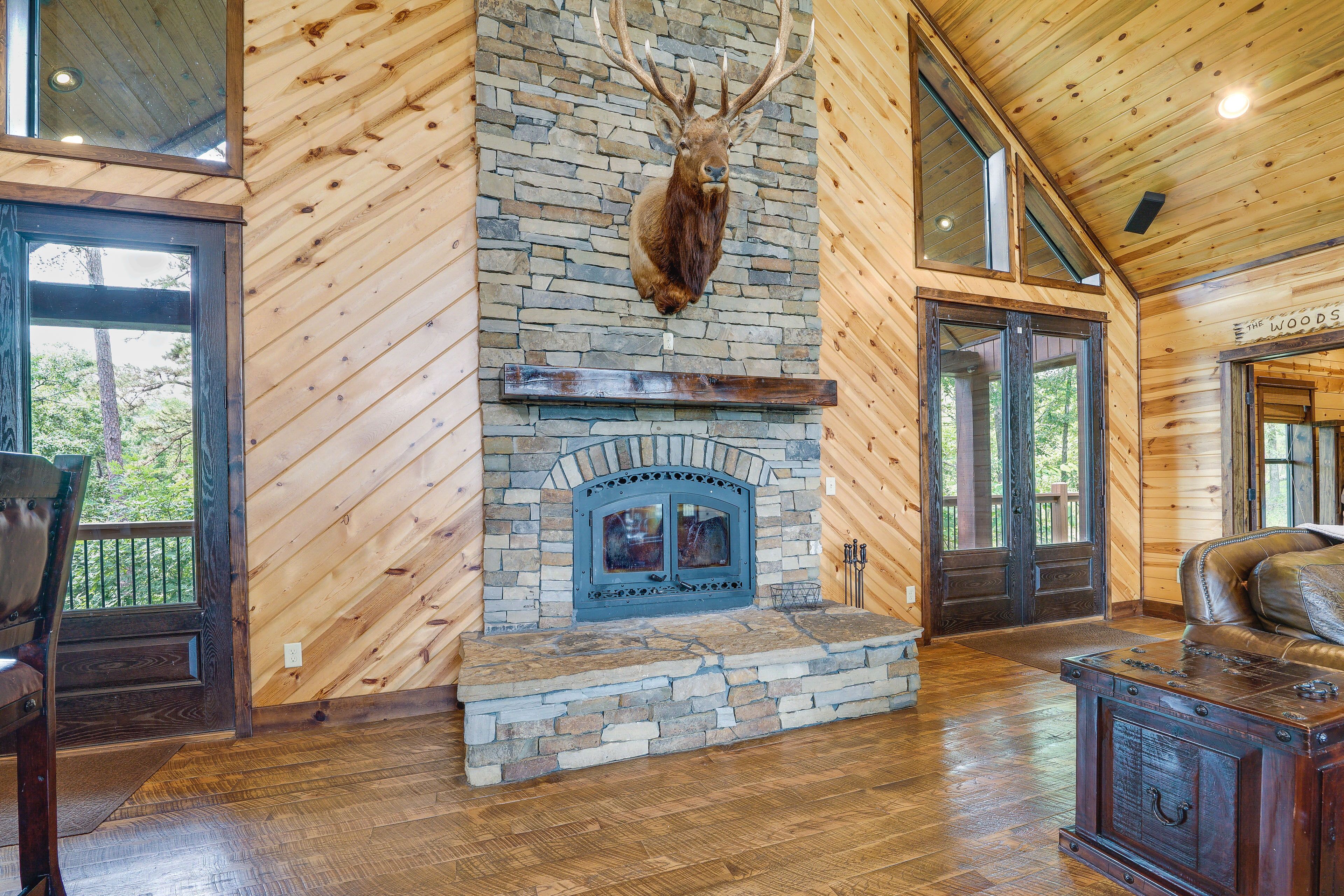 A log cabin interior with a stone fireplace, wooden walls, and a mounted deer head.