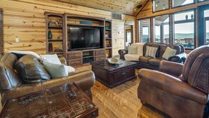 A cozy living room with wooden walls, leather sofas, a flat-screen TV, and large windows offering a view. - Broken Bow Cabin w/ Game Room & Private Hot Tub! (Broken Bow)