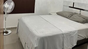 Ein Schlafzimmer mit einem Bett, einem Nachttisch, einem Wandventilator und einem Fenster mit Jalousie.