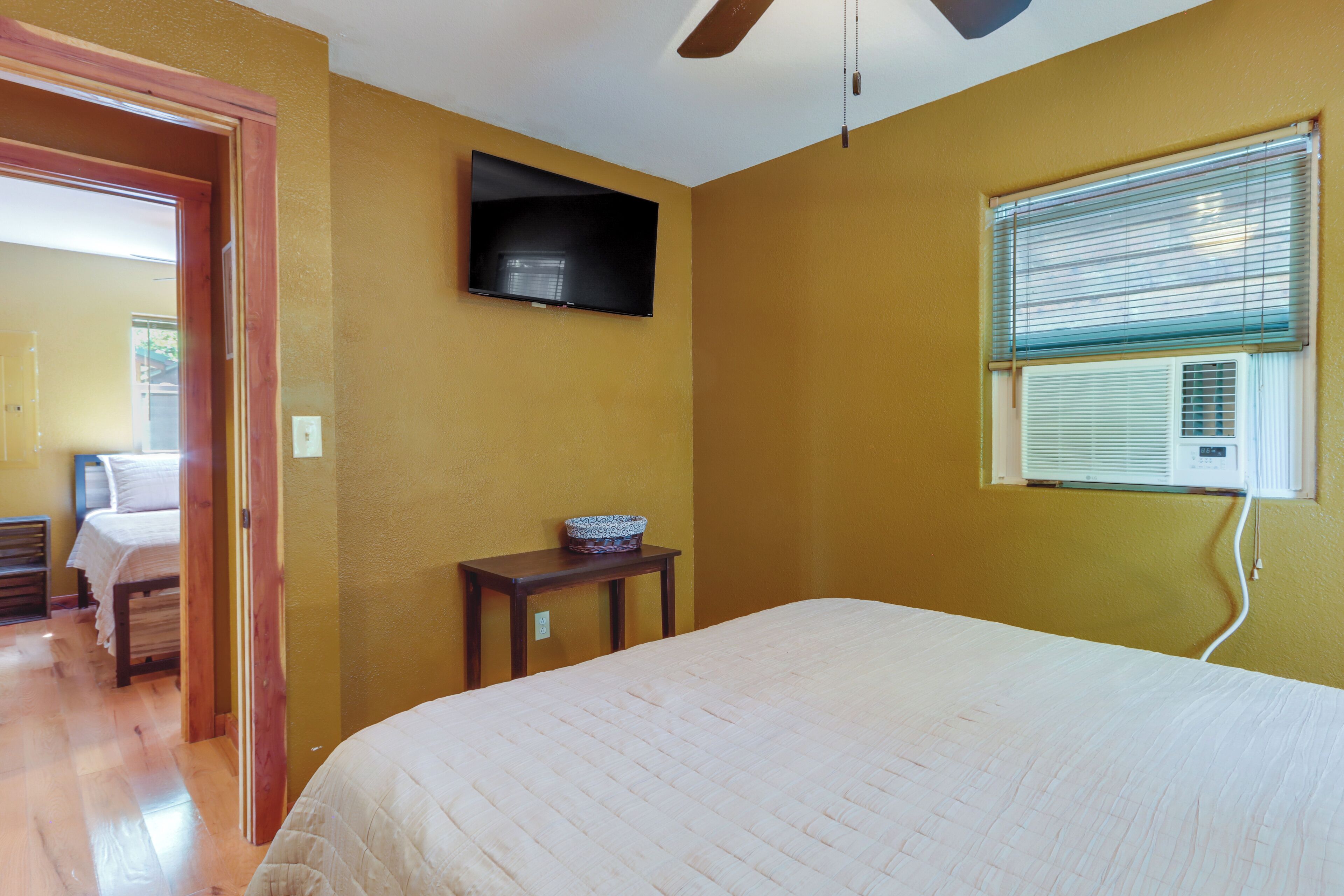 Un dormitorio con cama, televisor montado en la pared, ventana con persianas y una mesita con un frutero.