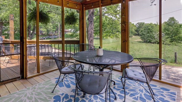 Eine überdachte Terrasse mit einem runden Tisch und Stühlen, umgeben von großen Fenstern mit Blick auf eine grüne Landschaft.
