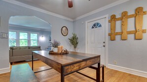 A spacious living room with a wooden dining table, a bench, and a sofa. There is a door with a wooden cross decoration, a clock, and a window with a view of greenery. - Vacation Rental Near University of Illinois (Champaign)
