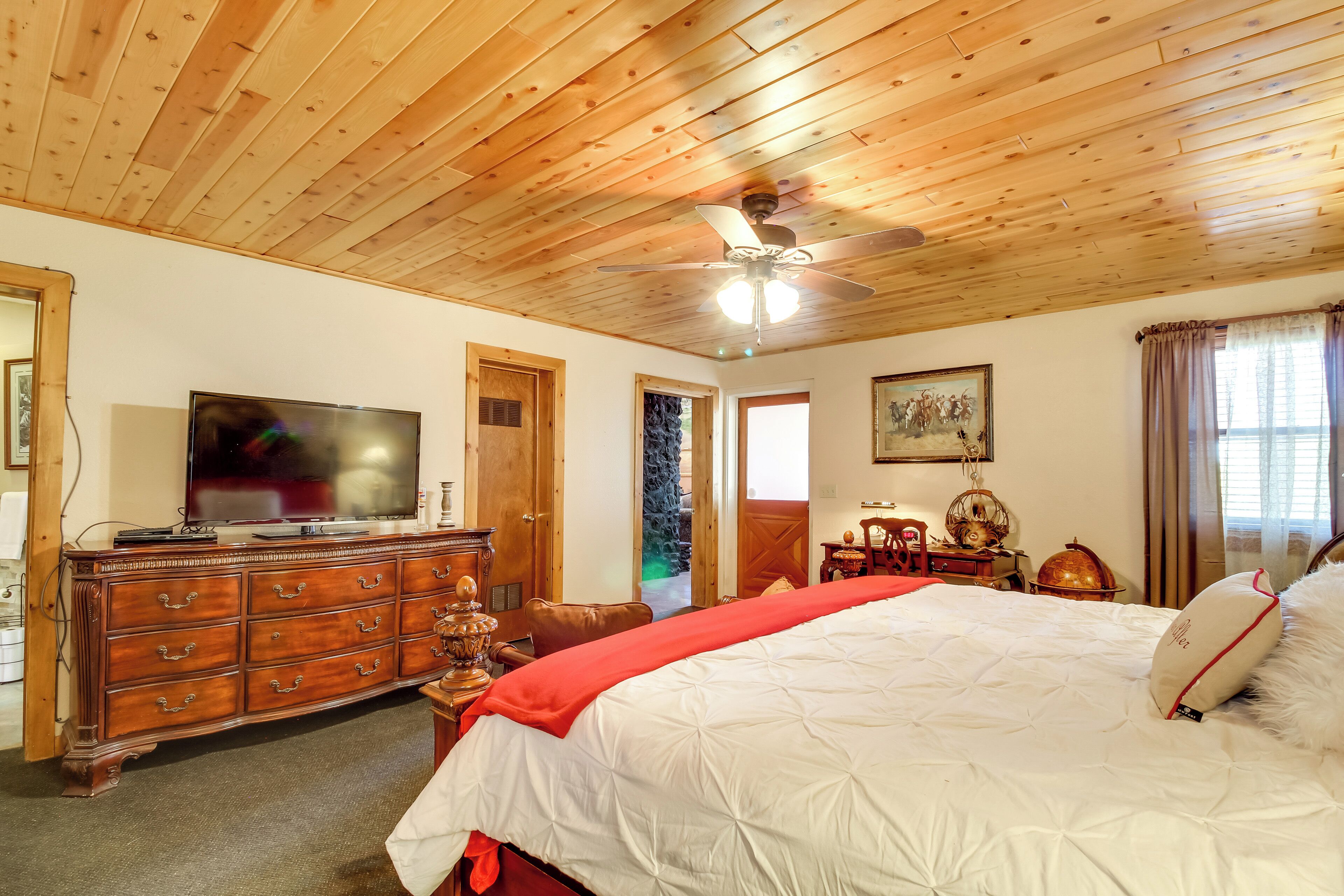 A bedroom with a wooden ceiling, a bed with white bedding, a wooden dresser, a television, and a ceiling fan.