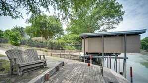 A wooden dock with a bench and a covered structure by a calm body of water, surrounded by trees and a fence.