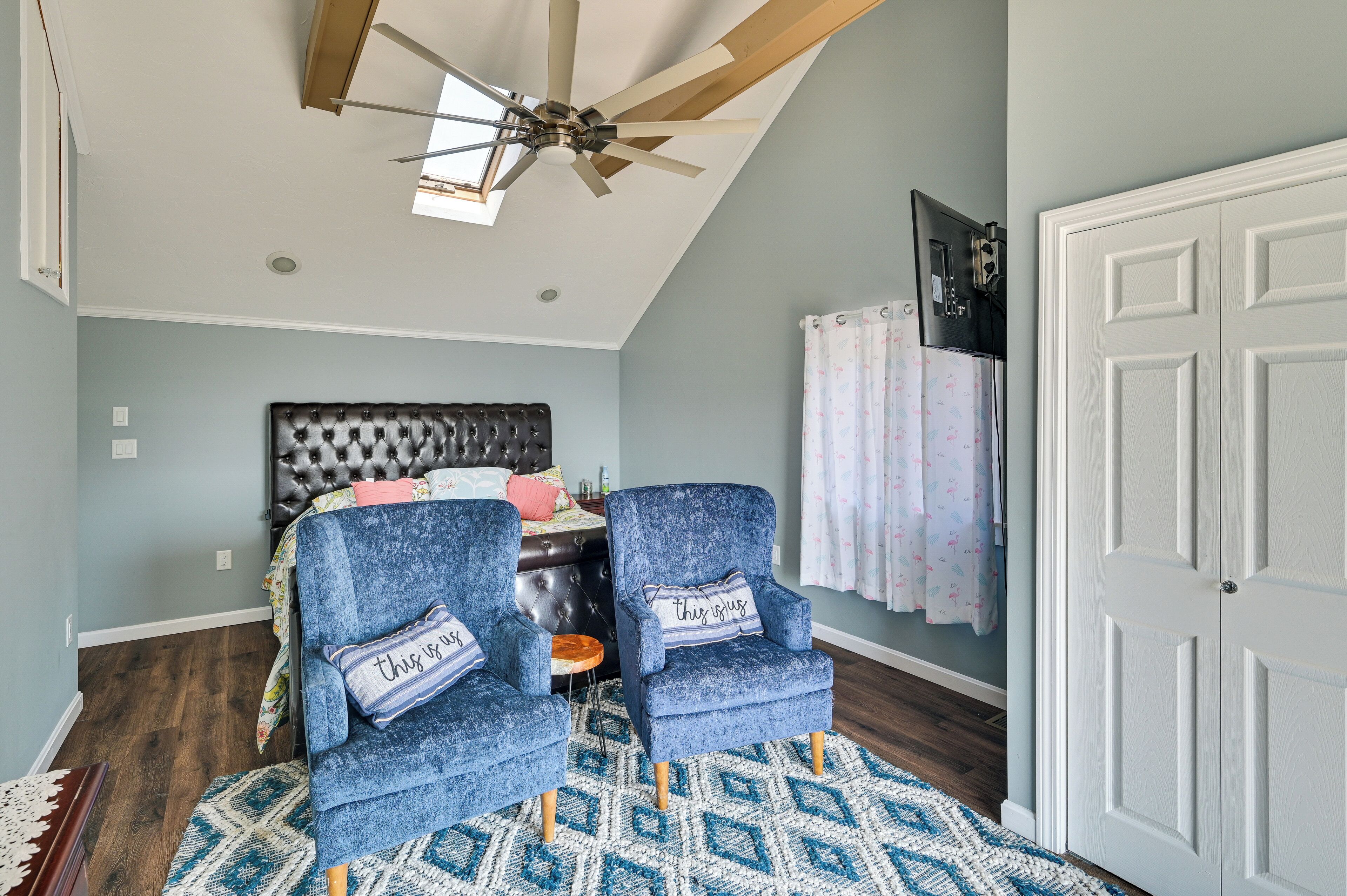 A bedroom with a bed, two armchairs, a ceiling fan, a patterned rug, and a white door.