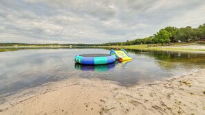 A calm lake with a sandy shore, a blue and yellow inflatable pool, and a grassy area with trees in the background. - Lakefront Keystone Heights Home w/ Boat Ramp! (Keystone Heights)