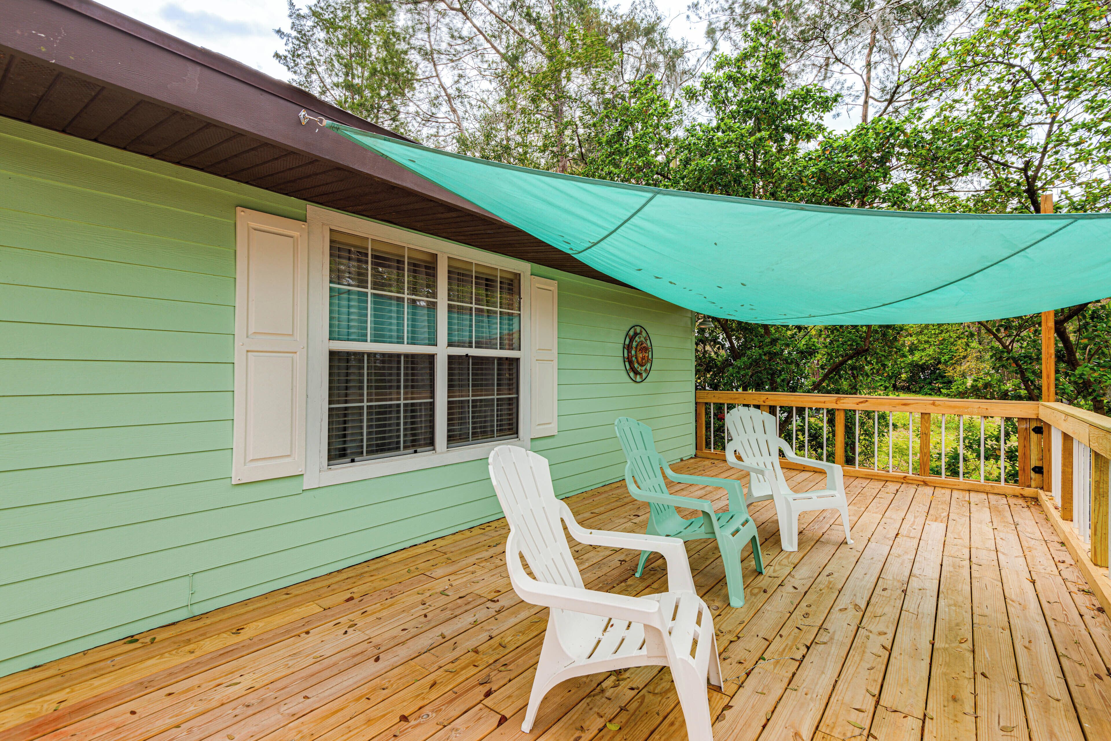 A wooden deck with two white Adirondack chairs and a table, a teal awning, and a window with white shutters.