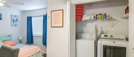 Un dormitorio compacto con cama, ventilador de techo y zona de lavandería con lavadora y tendedero.