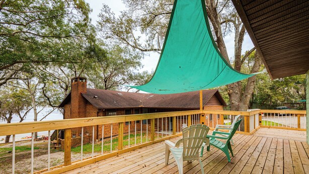 A wooden deck with a green chair and a table, overlooking a beach and a brick building. - Lakefront Keystone Heights Home w/ Boat Ramp! (Keystone Heights)