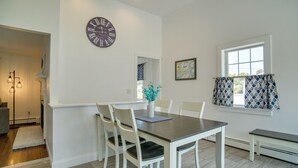 A dining area with a wooden table and white chairs, a wall clock, and a window with patterned curtains. - Newly Renovated Dennis Port Home: Close to Beaches (Dennis Port)