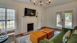 A living room with a fireplace, a television mounted above it, and a green sofa with a floral pillow. There are two wooden ottomans in front of the sofa, and a patterned rug on the floor. - Walk to Bergfeld Park: 1928 Azalea District Home (Tyler)
