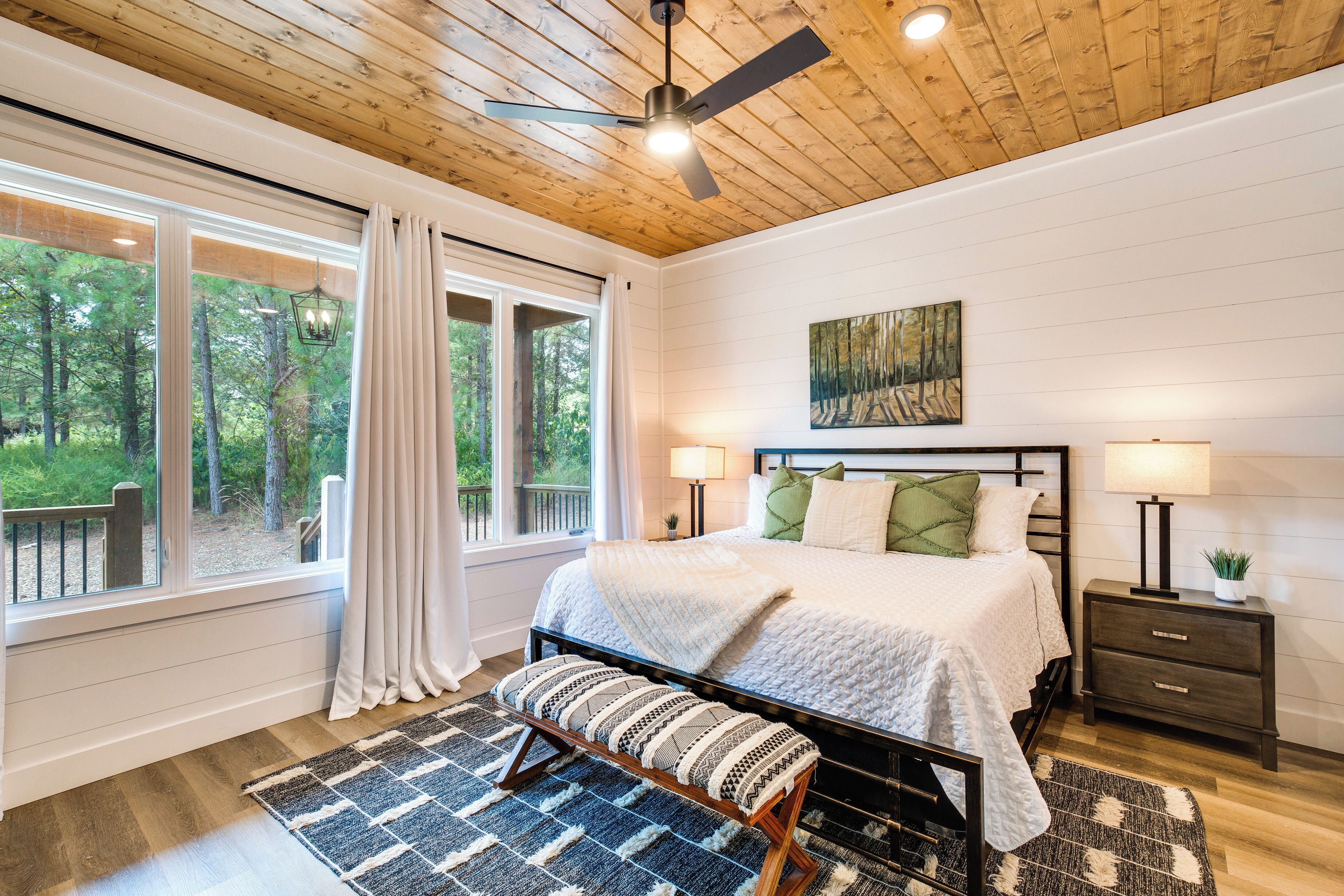 A bedroom with a bed, bedside tables, a bench, a ceiling fan, and a window with curtains.