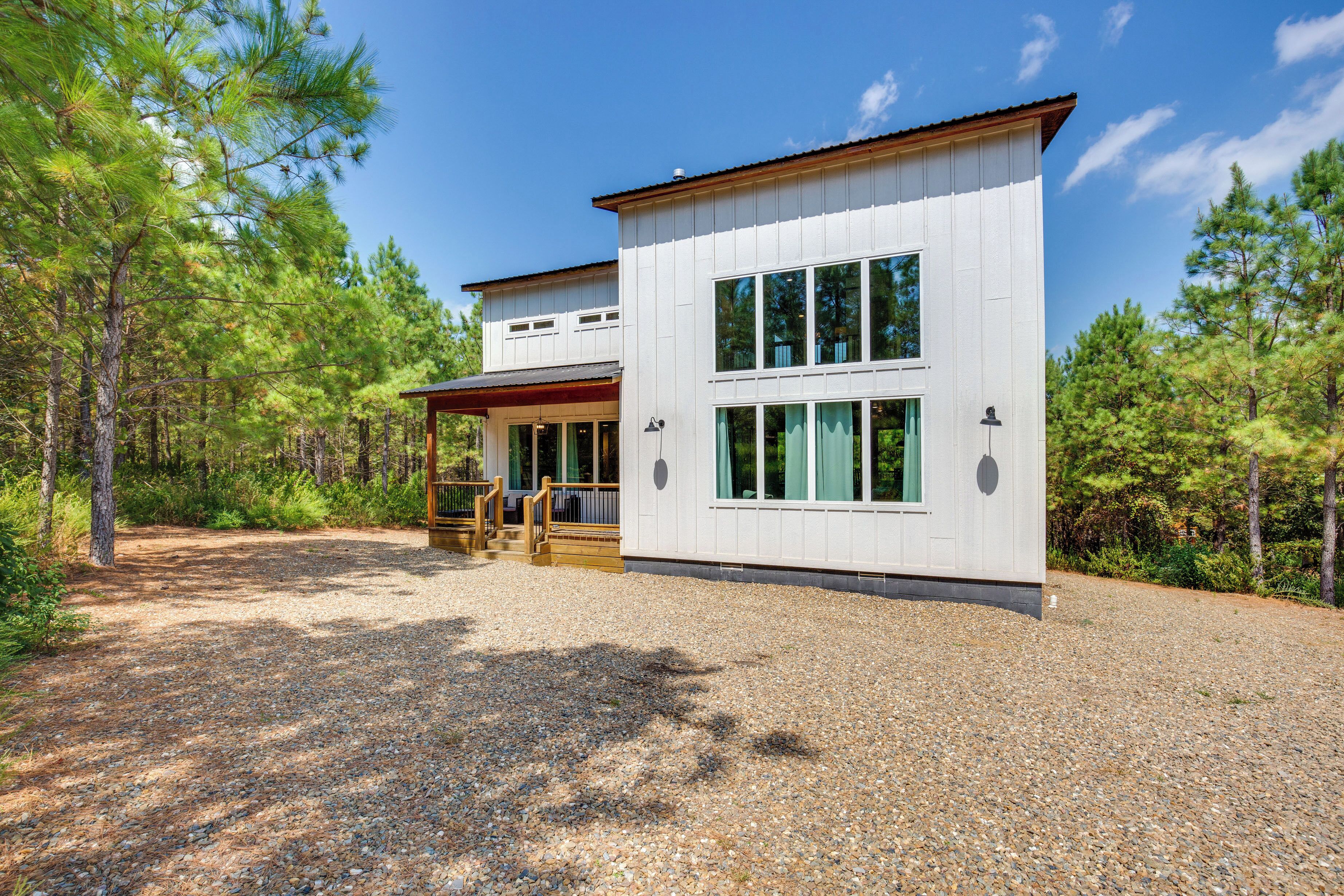 A modern two-story cabin with a gravel driveway and a wooden deck, surrounded by pine trees.