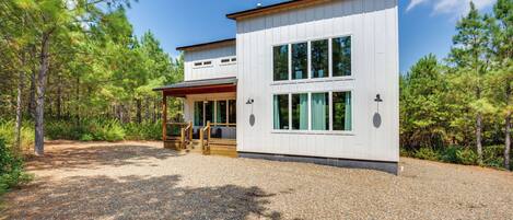 A modern two-story cabin with a gravel driveway and a wooden deck, surrounded by pine trees.