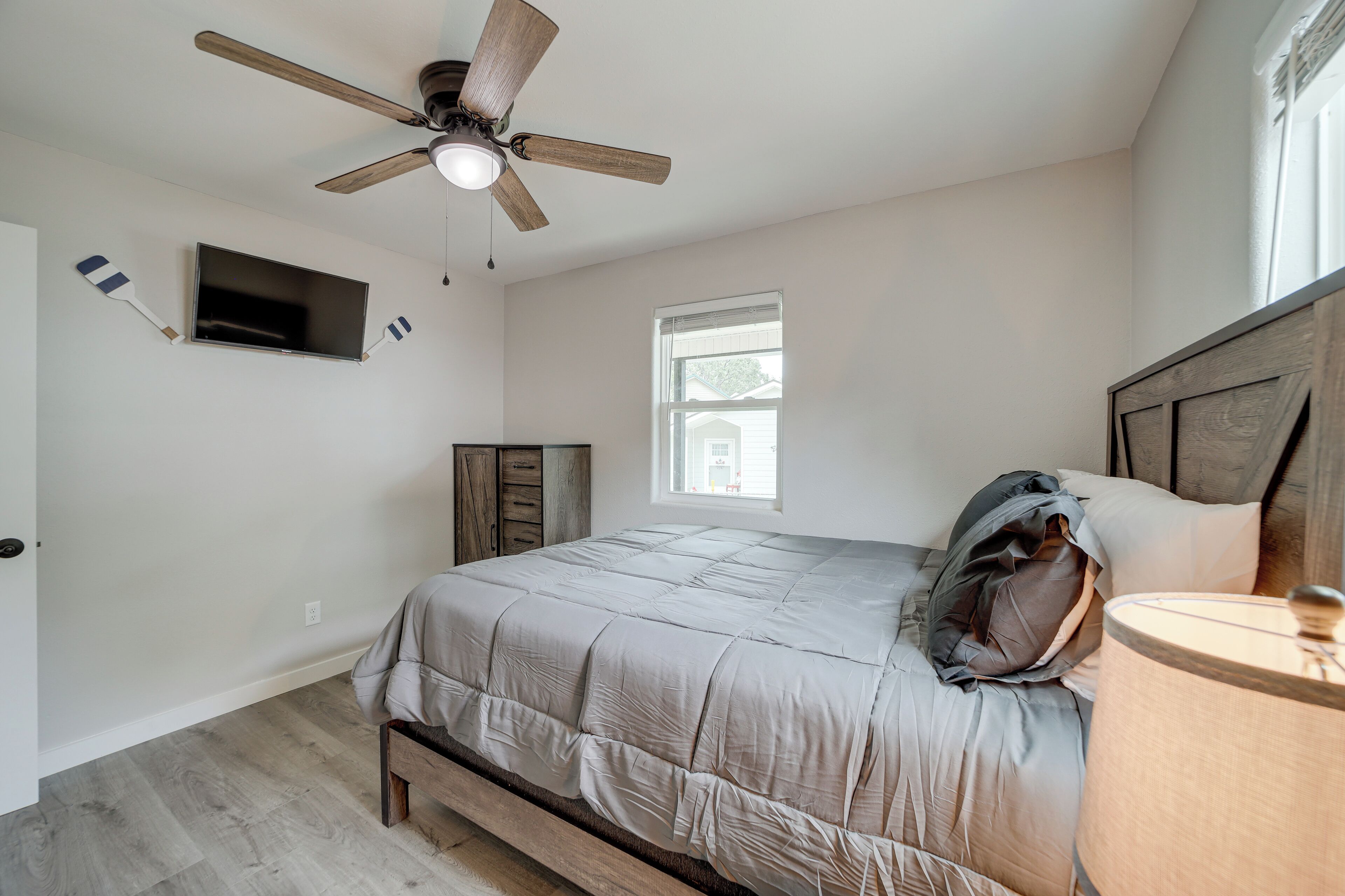 A bedroom with a bed, a ceiling fan, a TV mounted on the wall, a window, and a wooden headboard.